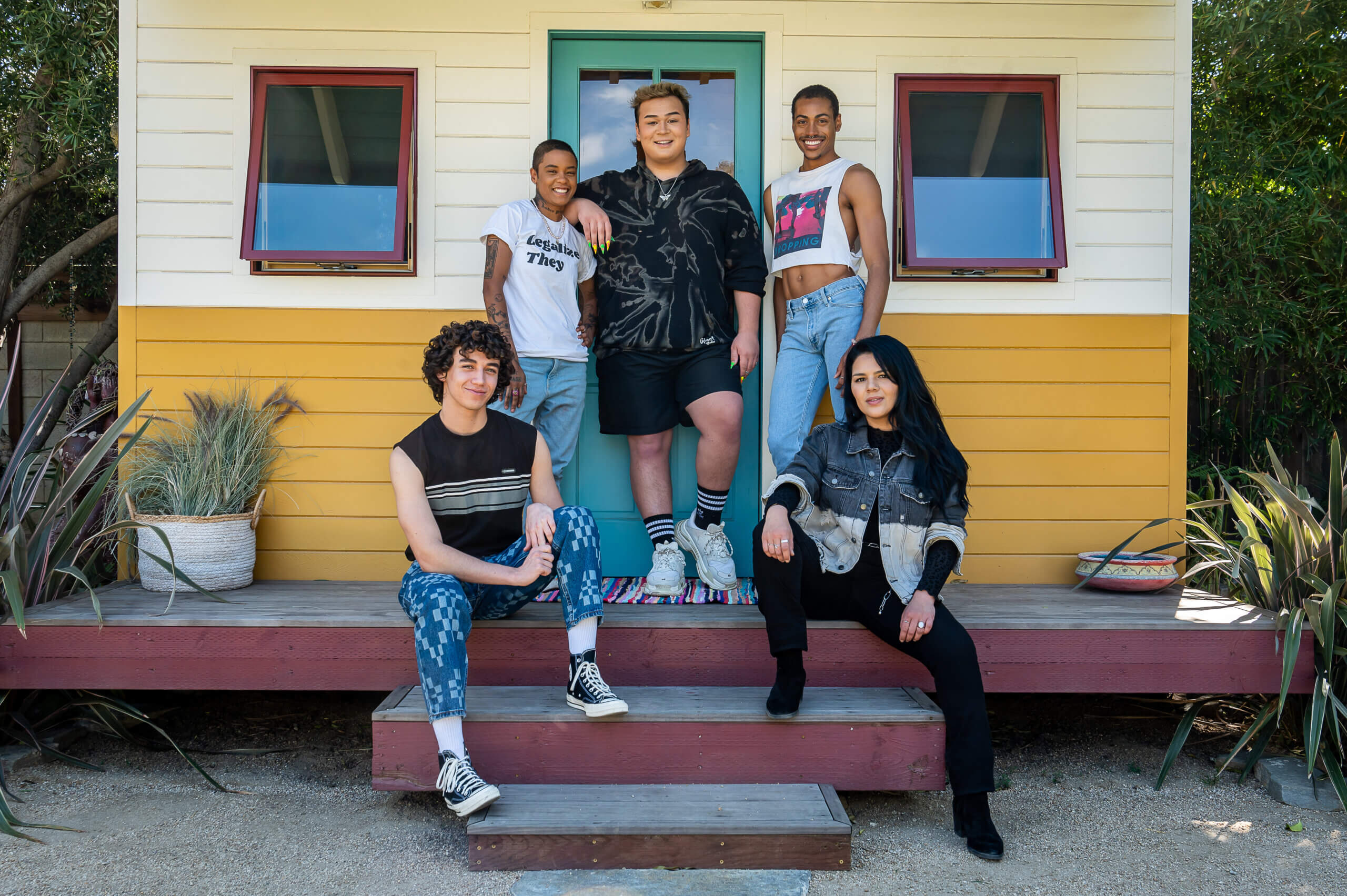 A diverse group of five LGBTQ+ young people smile in front of a small yellow beach house.
