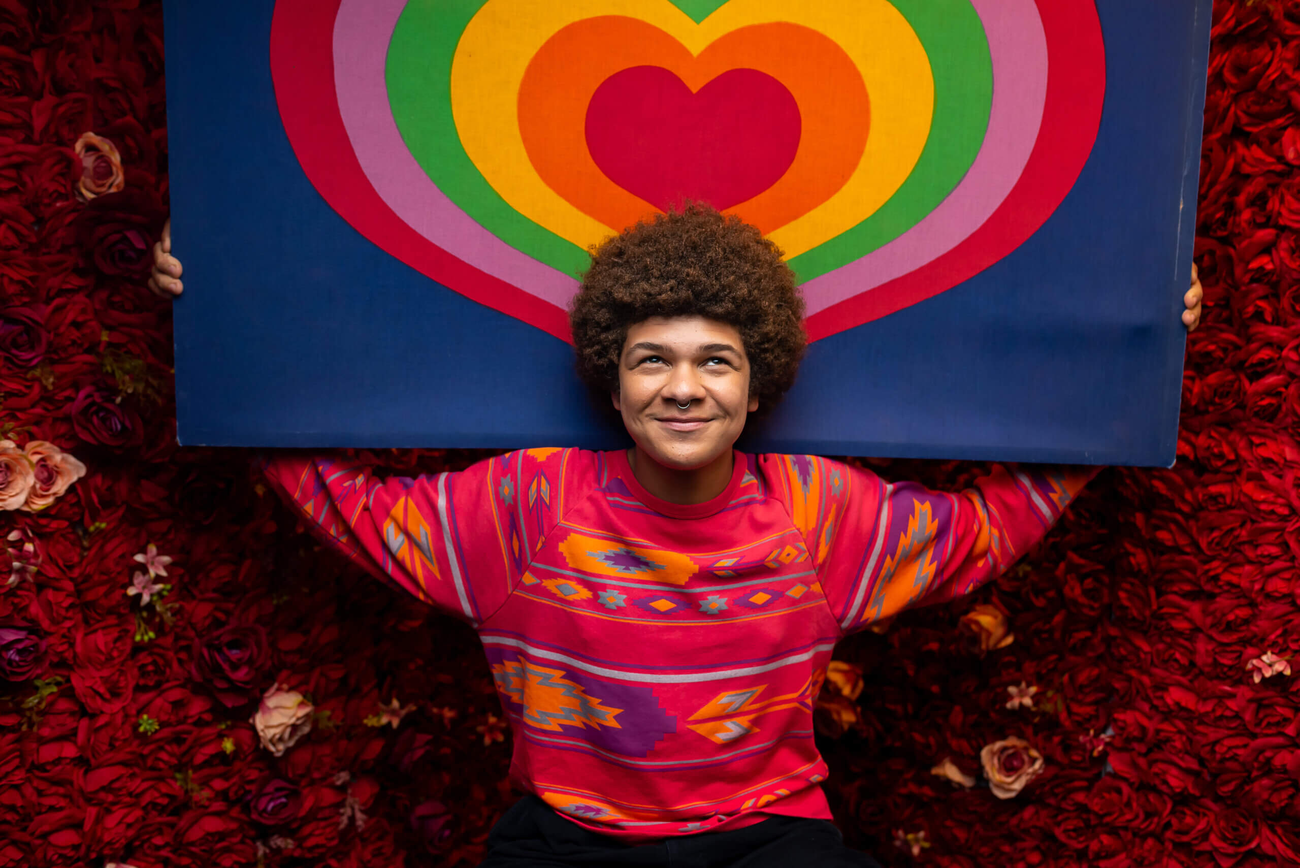 An LGBTQ+ young person smiles and holds a picture of a heart.